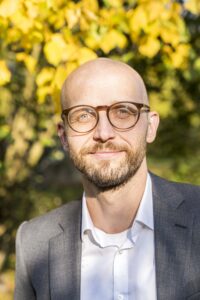 Gregor Herda in a suit in front of an autumnal leafy background.